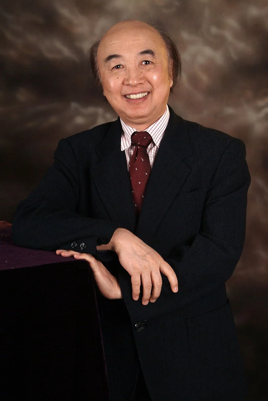 Portrait of a smiling man in a black suit with a red tie, leaning on a surface with his right arm, against a brown textured background.