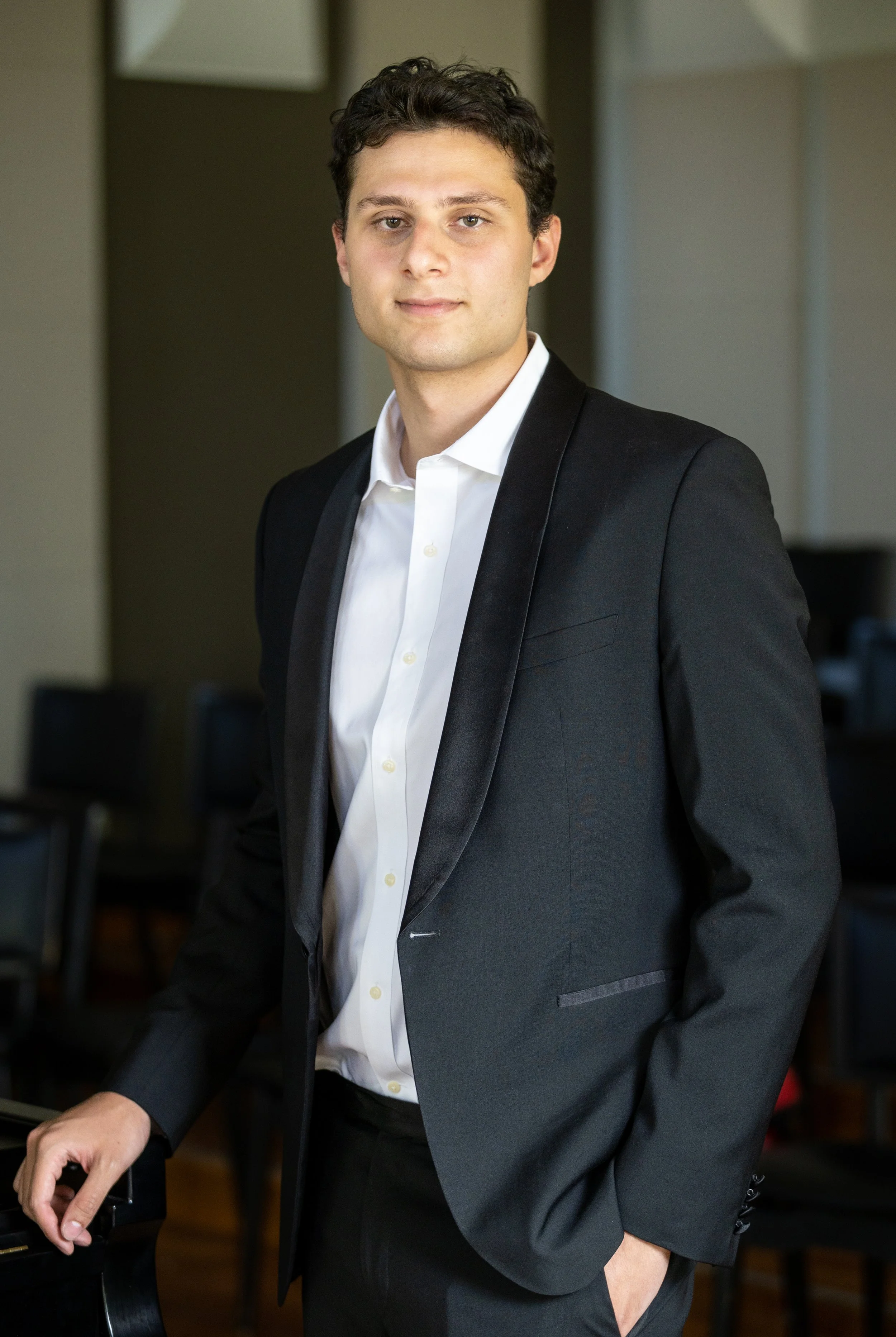 A young man in a black suit with arms crossed, standing in front of a blurred background with a banner and wall art.