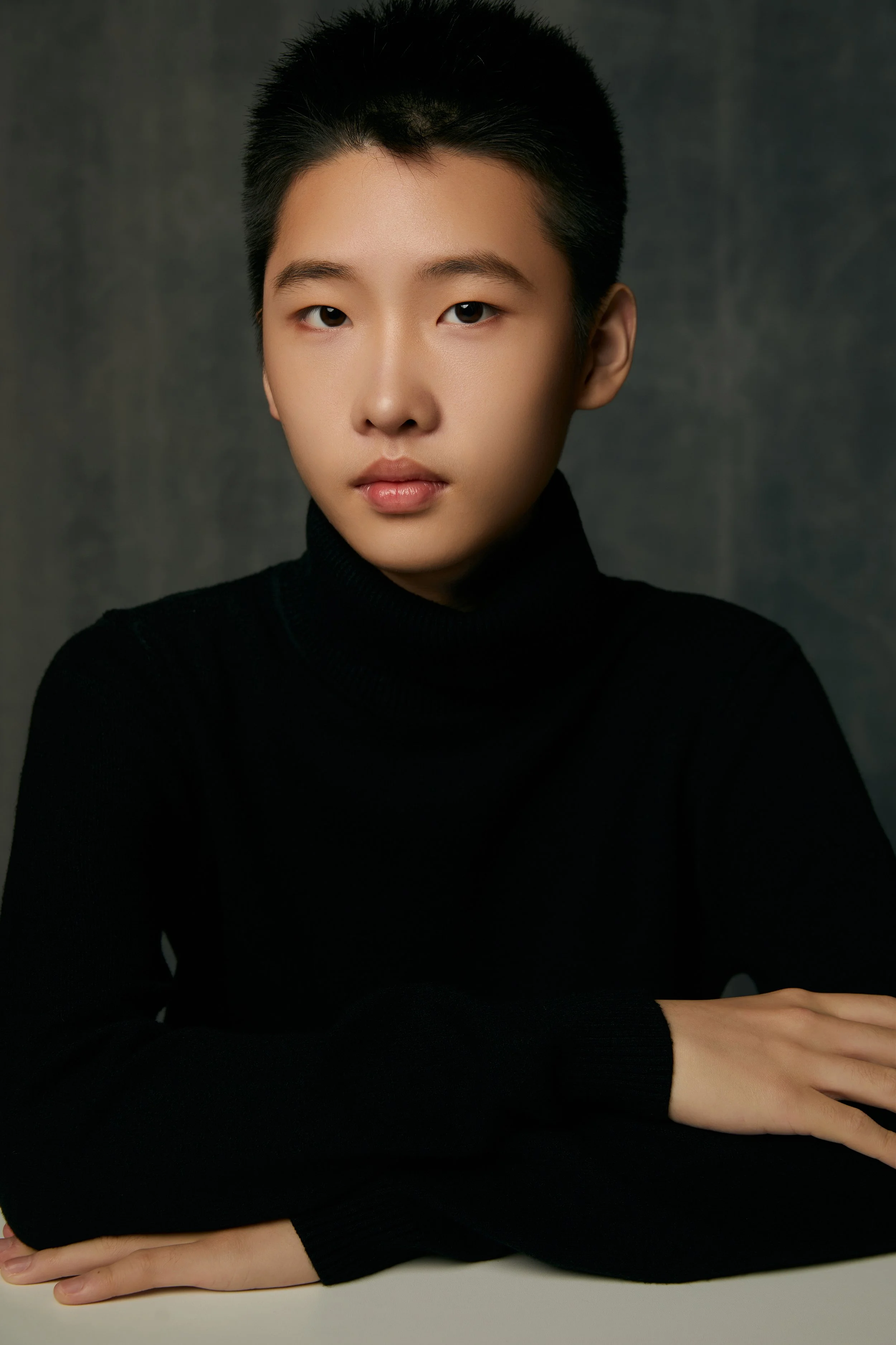 A young woman in a black sleeveless dress stands with her arms crossed, smiling softly at the camera, in an indoor setting with a blurred background and a banner behind her.