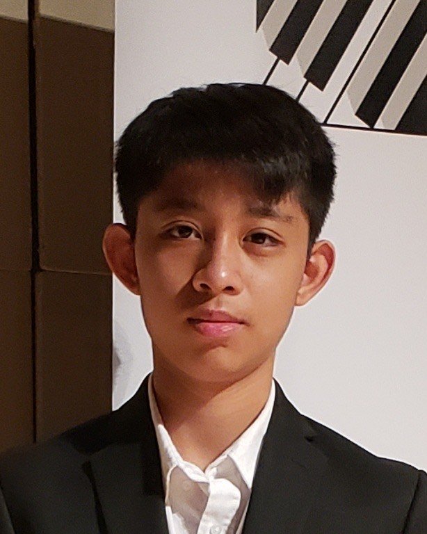 A young man in a black suit with arms crossed, standing in front of a blurred background with a banner and wall art.