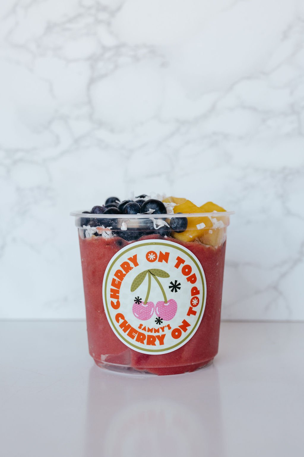 Fruit smoothie bowl with blueberries, mango, and shredded coconut in a clear cup labeled "Cherry On Top."