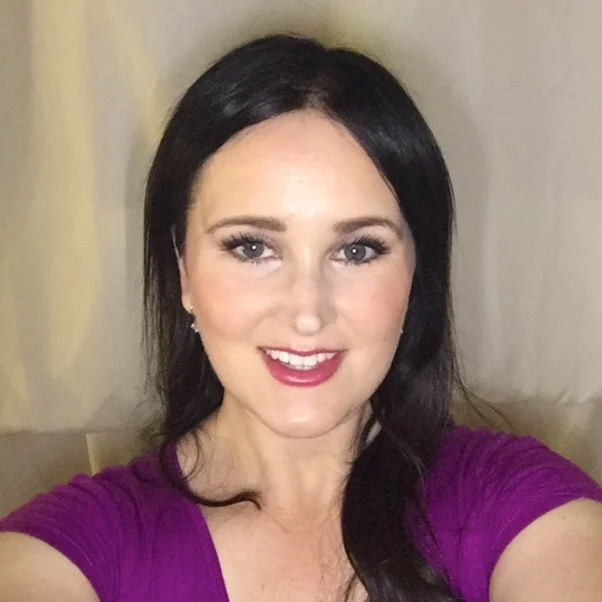 A woman with long dark hair, wearing a purple top, smiling at the camera indoors.
