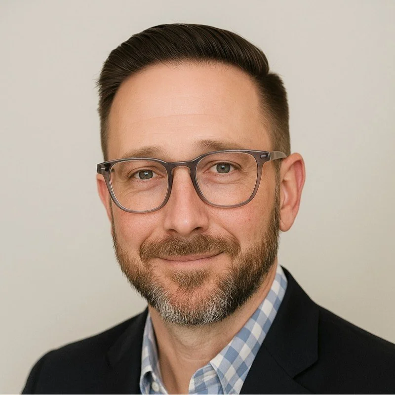 Close-up of a middle-aged man with glasses, a beard, wearing a checked shirt and dark blazer, smiling against a plain background.