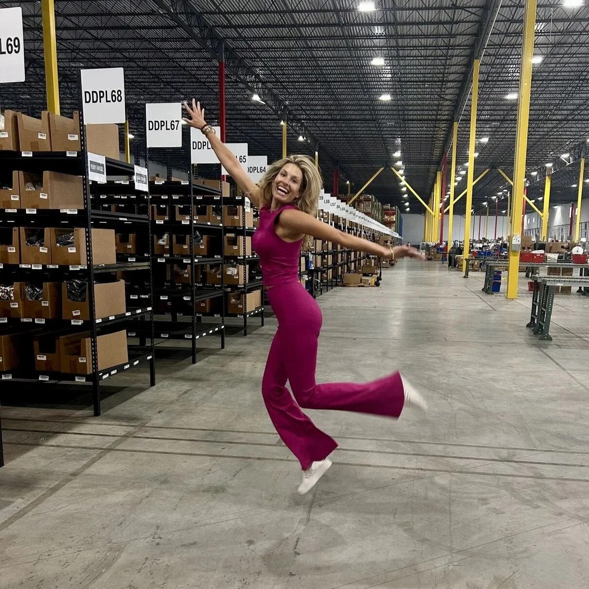 Woman in a magenta jumpsuit joyfully jumping in a warehouse aisle with shelves and boxes in the background.