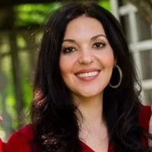 A woman with dark hair smiling, wearing hoop earrings and a red top, outdoors with greenery and a house in the background.