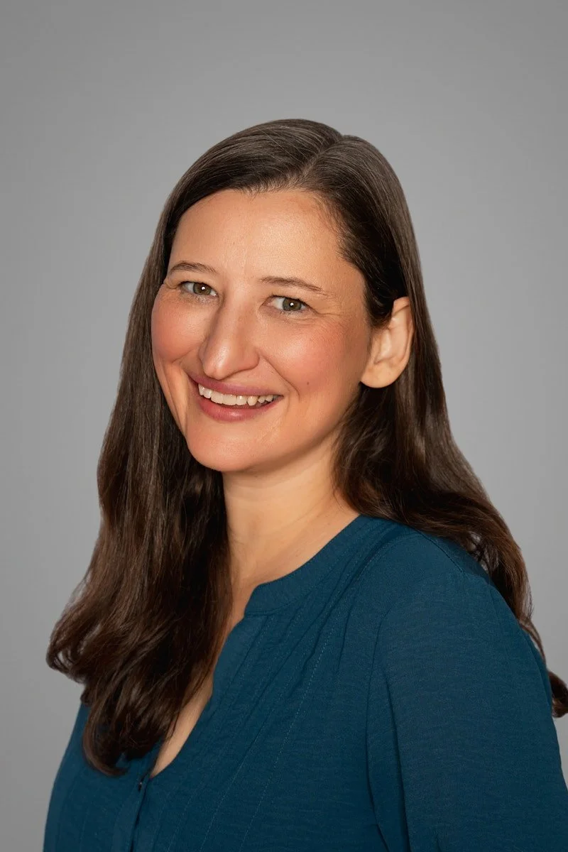 The owning clinician of the woman-owned business with long brown hair, smiling, wearing a blue top, against a gray background.