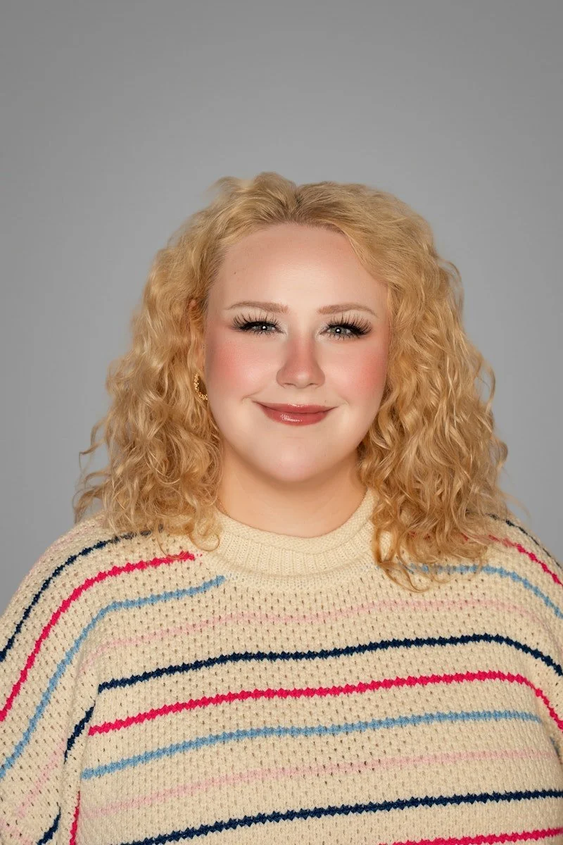 Rachel Brown, MSW, LSW, a queer therapist in Cincinnati, OH, with blonde curly hair wearing a multicolored striped sweater and gold earrings, smiling against a plain gray background.