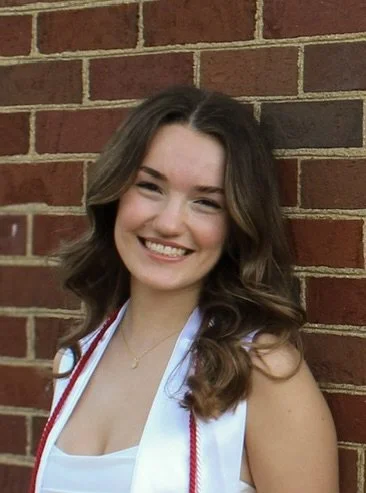 A woman neurodivergent-affirming with wavy brown hair smiling, wearing a white top and graduation stole, standing against a brick wall.