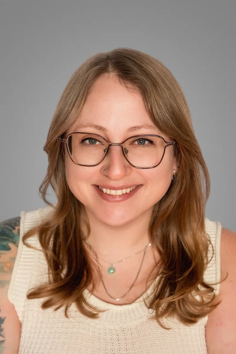 A female neurodivergent-affirming therapist in Cincinnati, OH, with light brown hair, wearing glasses, a nose piercing, and layered necklaces smiling at the camera against a solid gray background.
