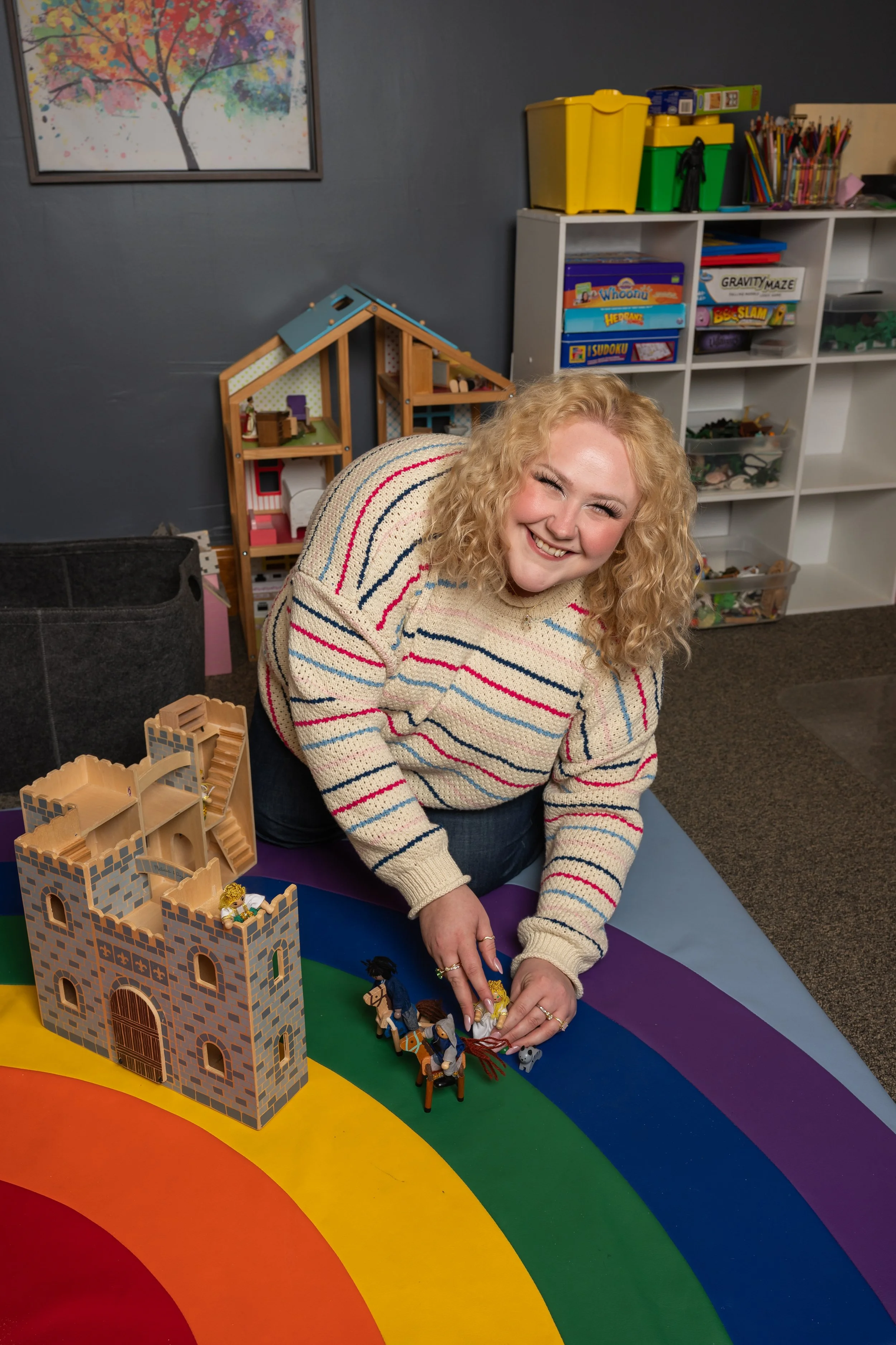 A smiling woman with curly blonde hair placing small toy figures on a rainbow-colored play mat. In the background, there is a bookshelf with games and toys, and a colorful painting of a tree on the wall.