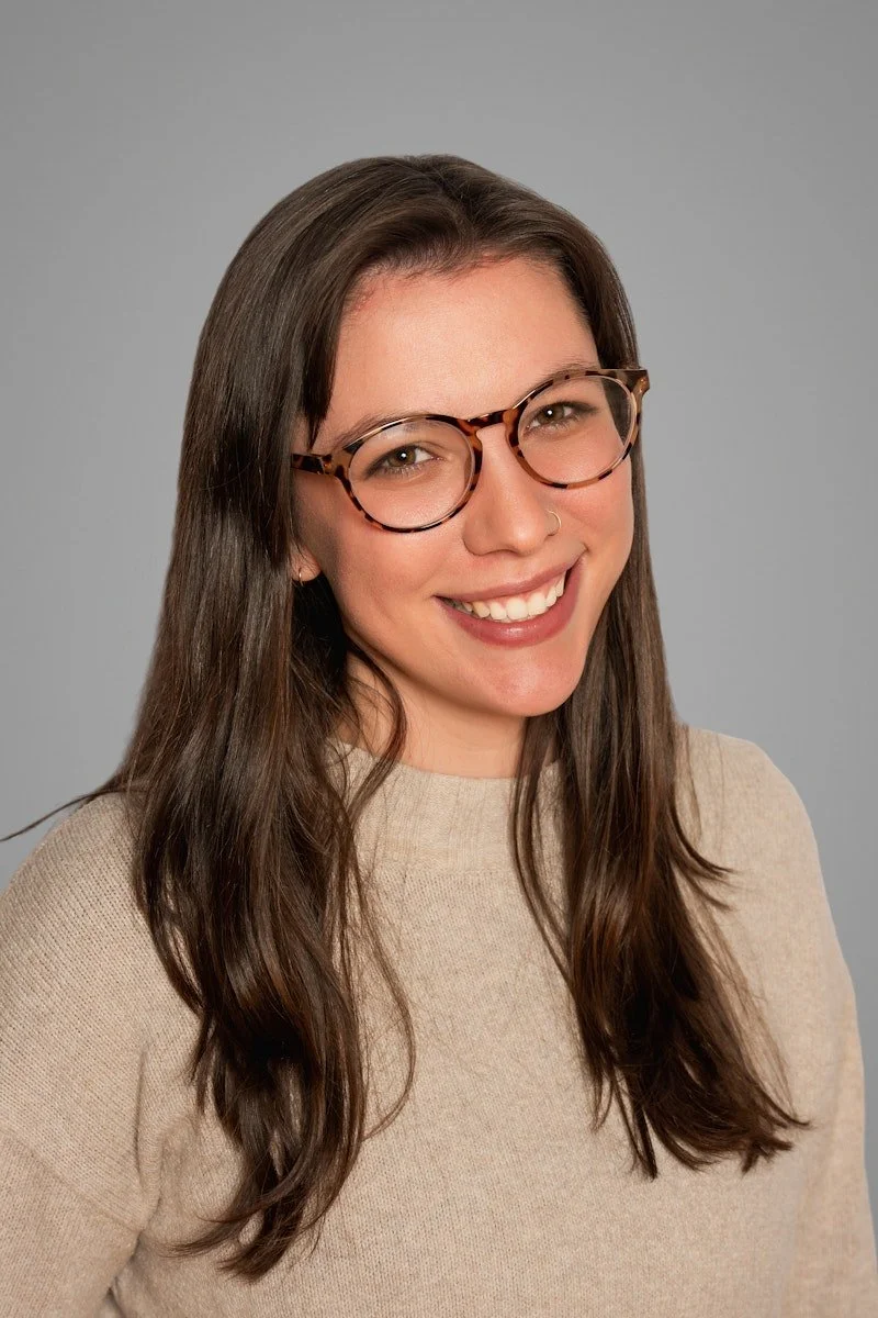 A smiling woman neurodivergent-affirming therapist in Cincinnati, OH, with long brown hair, wearing glasses with tortoise shell frames and a beige sweater, against a plain grey background.
