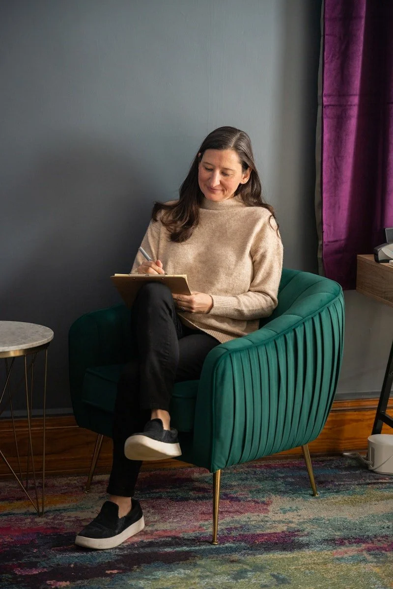 Dr. Elder, a Neurodivergent-affirming therapist in Cincinnati OH sitting in a green armchair, writing on a notepad with a pen smiling, in a room with a dark gray wall