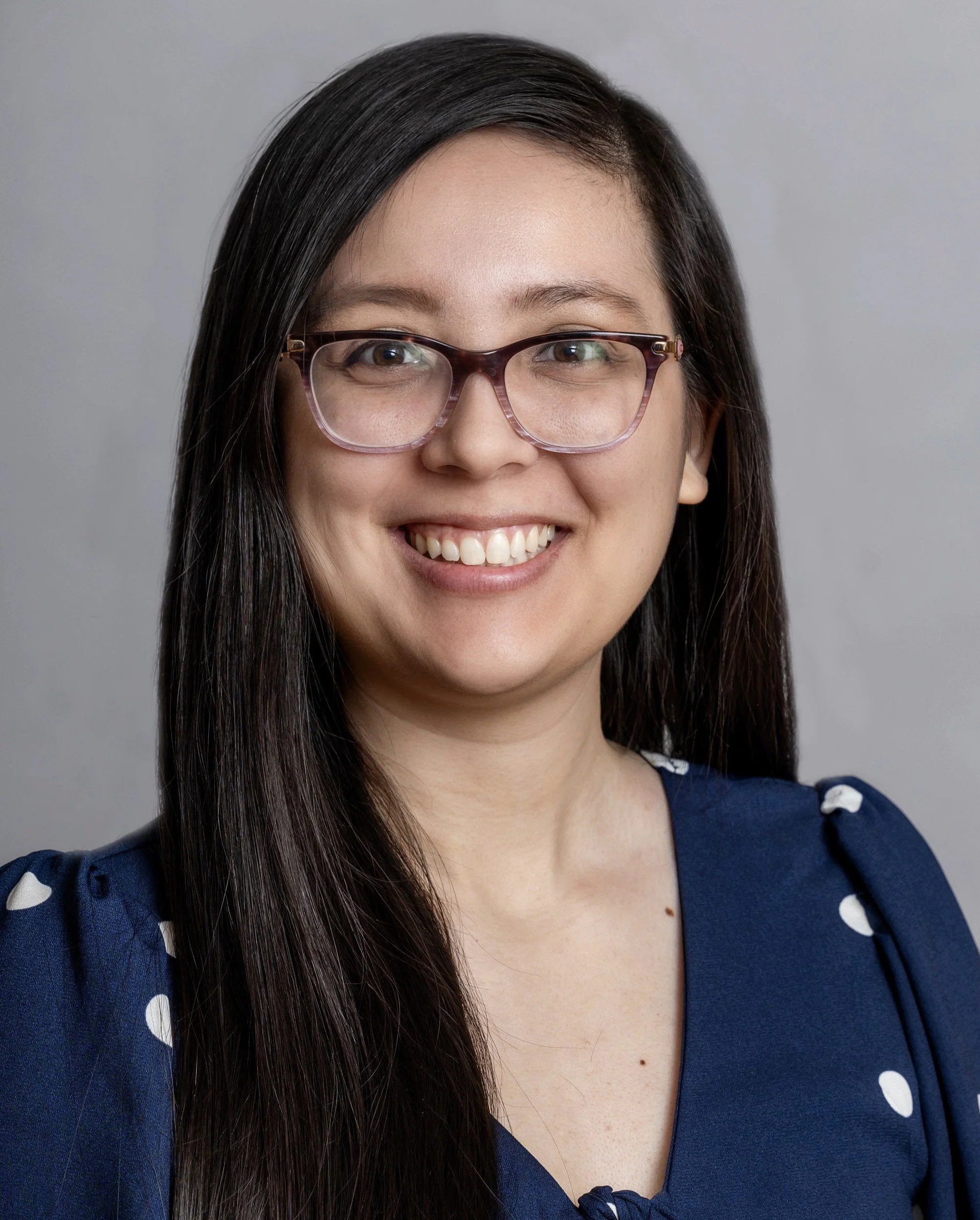 A smiling queer and nonbinary neurodivergent-affirming social worker  in Cincinnati, OH, with long dark hair, wearing glasses and a blue dress with white polka dots, against a neutral background.