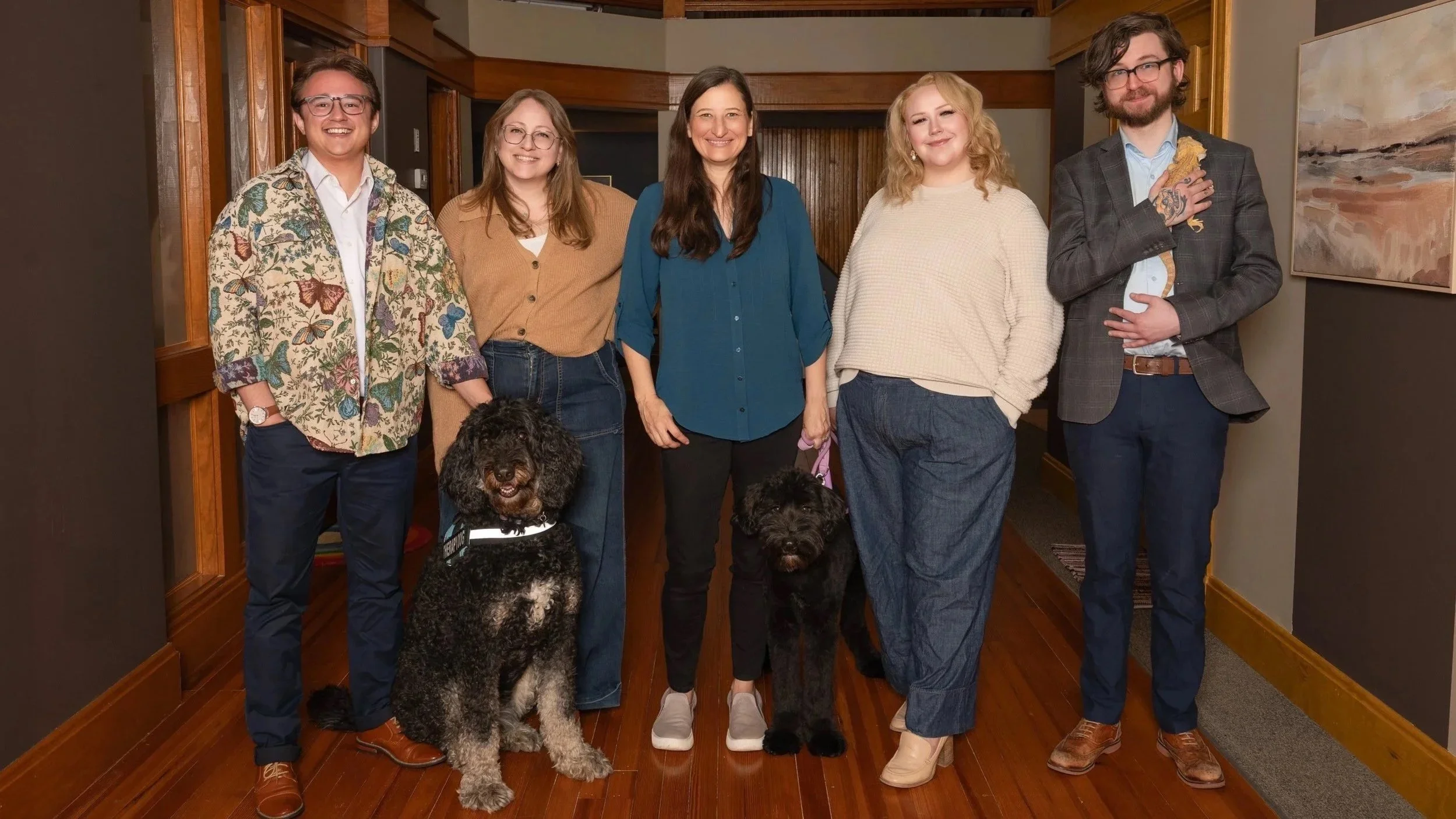 Group of six people, the team, standing indoors, smiling at the camera, with two dogs on leashes in front of them, and artwork on the wall to the right.