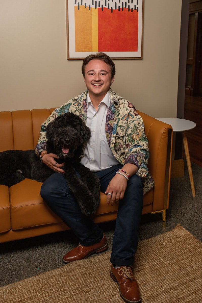 A young man sitting on a brown leather couch, smiling, holding a black dog with curly fur on his lap. Behind them is a wall with abstract artwork featuring orange, yellow, and black colors.