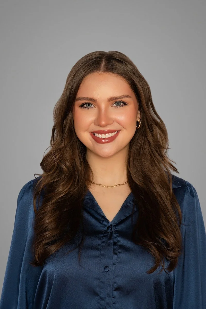 A woman neurodivergent-affirming therapist in Cincinnati, OH, with long, wavy brown hair and blue eyes, wearing a dark blue satin blouse, gold hoop earrings, a gold chain necklace, and smiling at the camera against a plain gray background.