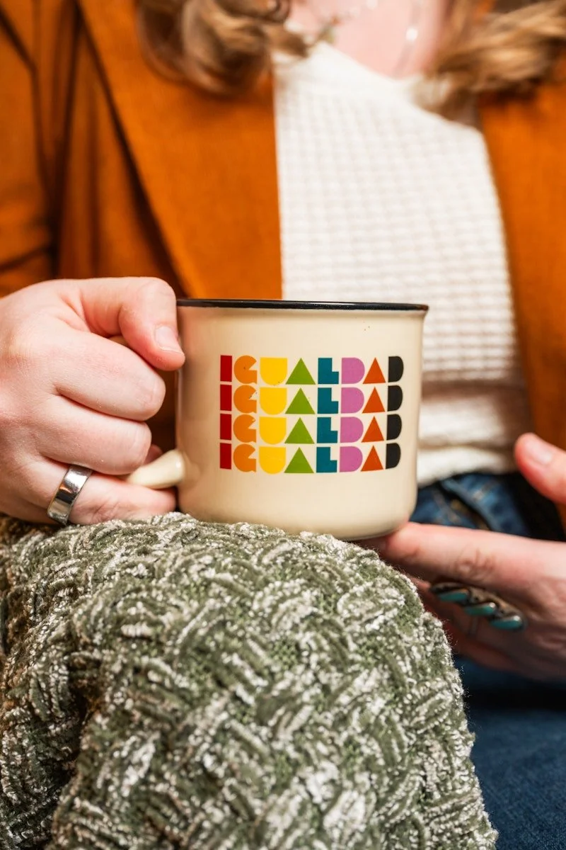 A person holding an enamel mug with the word "IGUALDAD" written in colorful, bold, and stylized letters. The person's hand is visible, wearing a ring, and they are sitting on a textured, green, fuzzy blanket. The background shows a woman wearing a brown jacket and a white, checkered shirt.