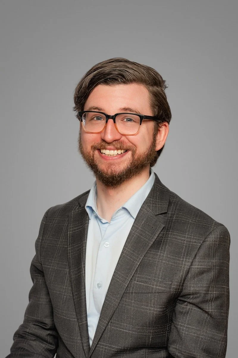 The male Ascent Psychological Services practice manager, in Cincinnati, OH, with glasses, a beard, and brown hair, wearing a dark plaid blazer and a light blue shirt, posing against a gray background.