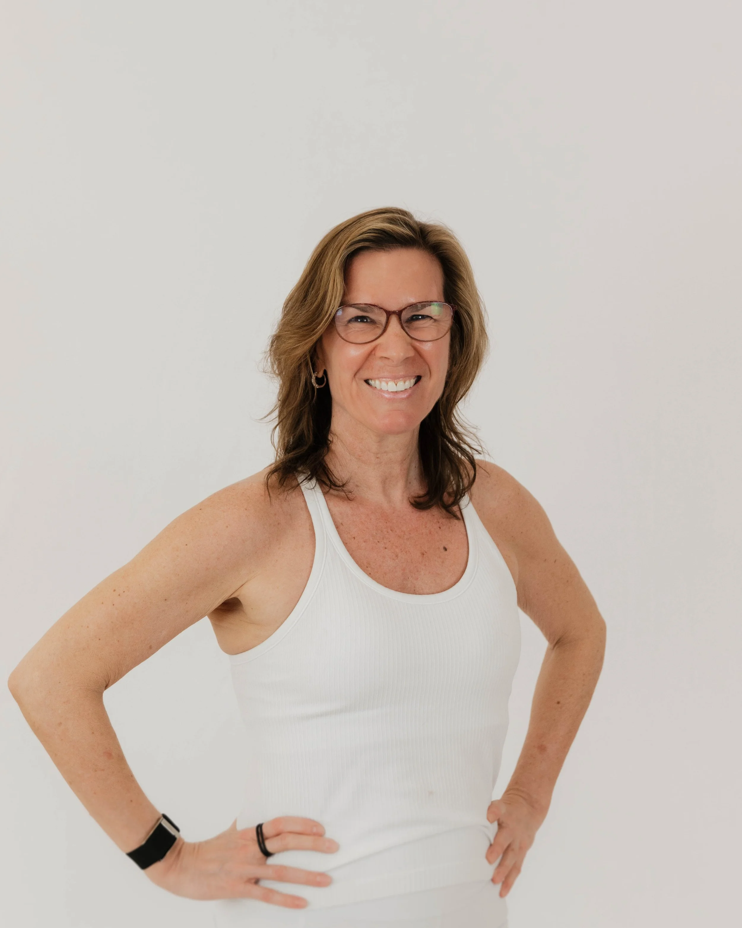 A smiling woman with short light brown hair, glasses, wearing a white tank top, against a light blue background.