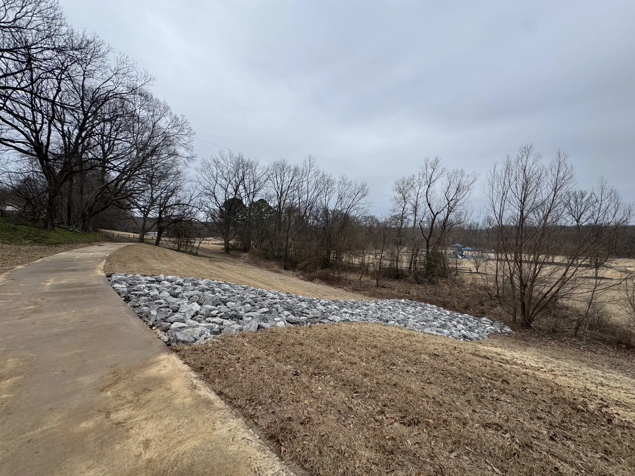 Empty park pathway with a curved concrete trail and a section of large gray rocks, surrounded by leafless trees and grassy areas, under an overcast sky.