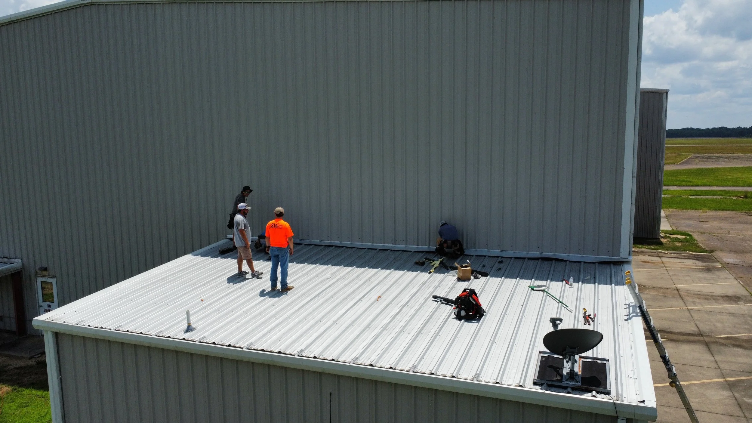 Four workers on a white metal roof of a large industrial building, installing or repairing the roofing with tools and equipment nearby, with a grassy field and cloudy sky in the background.