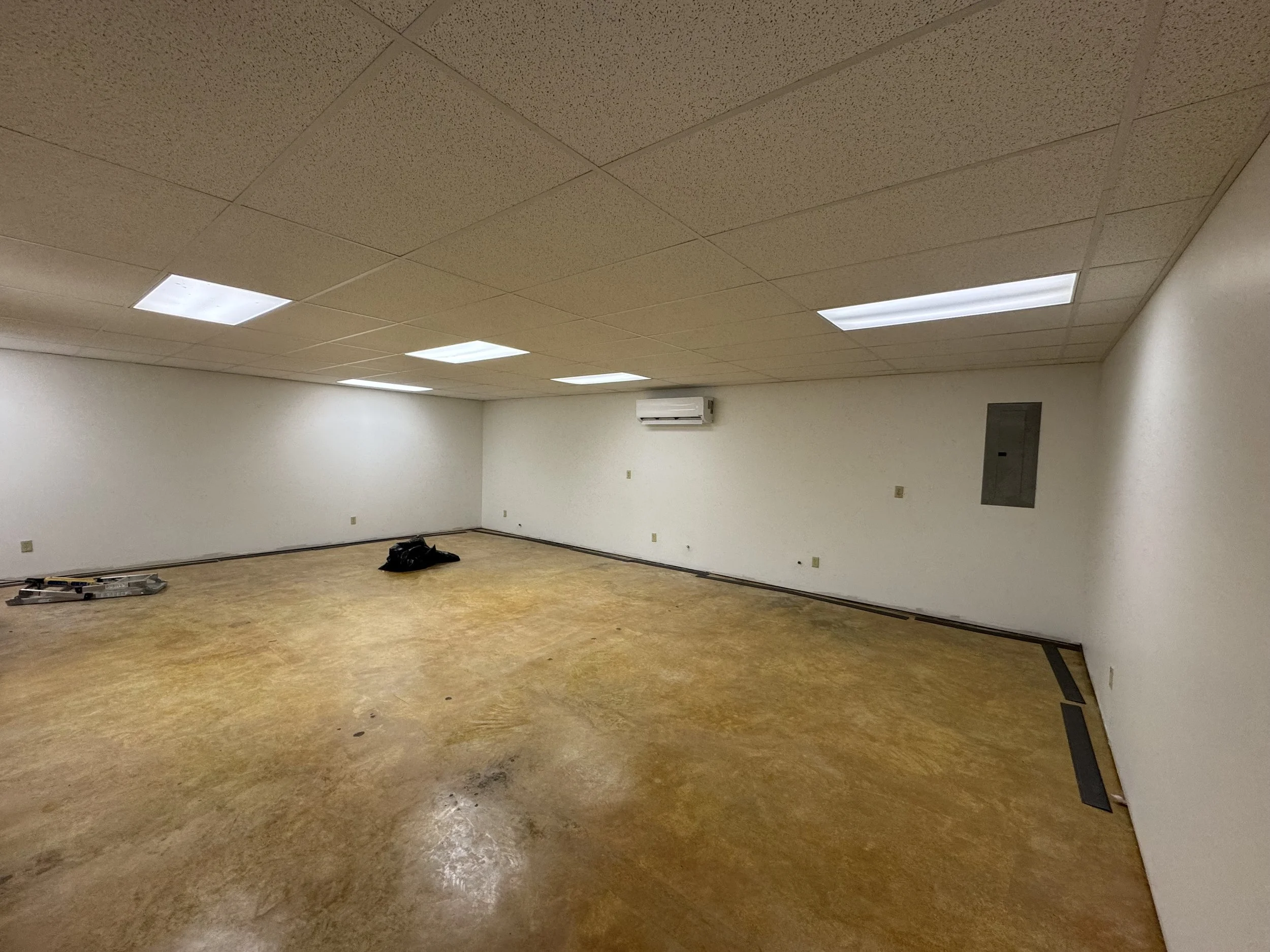 Empty office room with white walls, beige ceiling tiles, fluorescent lighting, and brown floor, with some tools and a black object on the floor.
