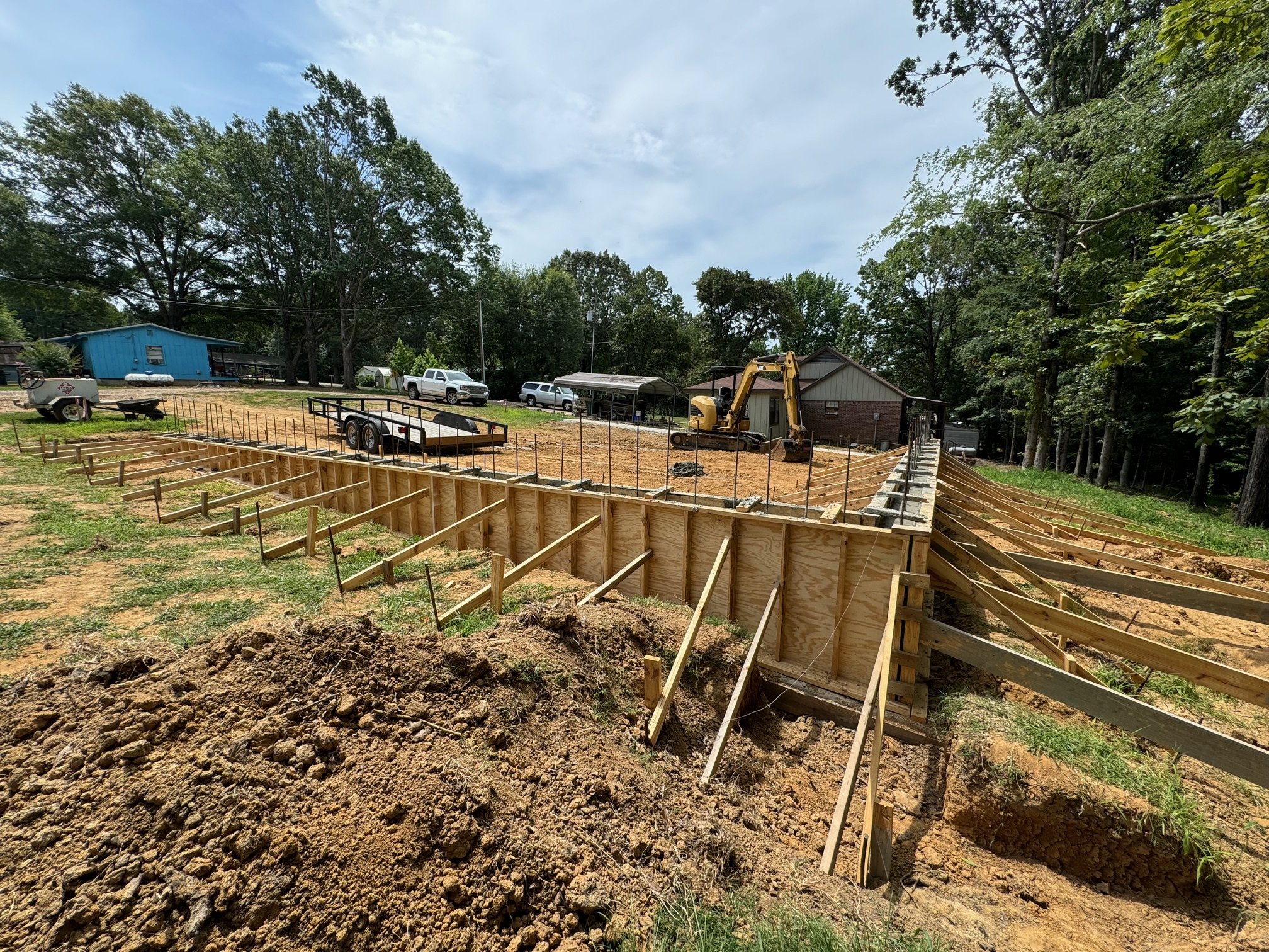 Construction site with wooden formwork for a concrete structure, a small excavator, parked trucks, and a grassy yard bordered by trees.