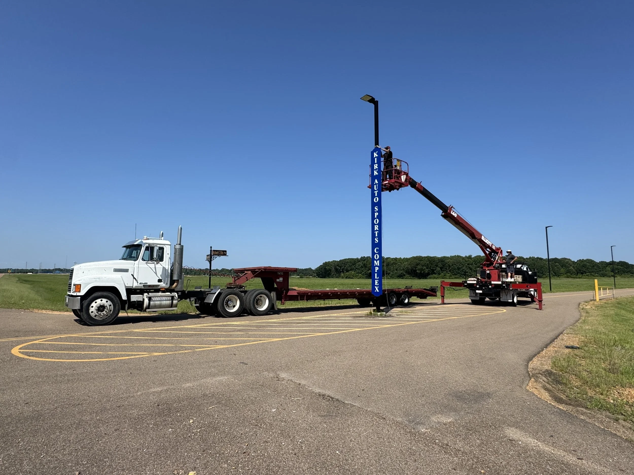 A truck with a cherry picker lift extending to a tall sports complex signpost in a parking lot under a clear blue sky.