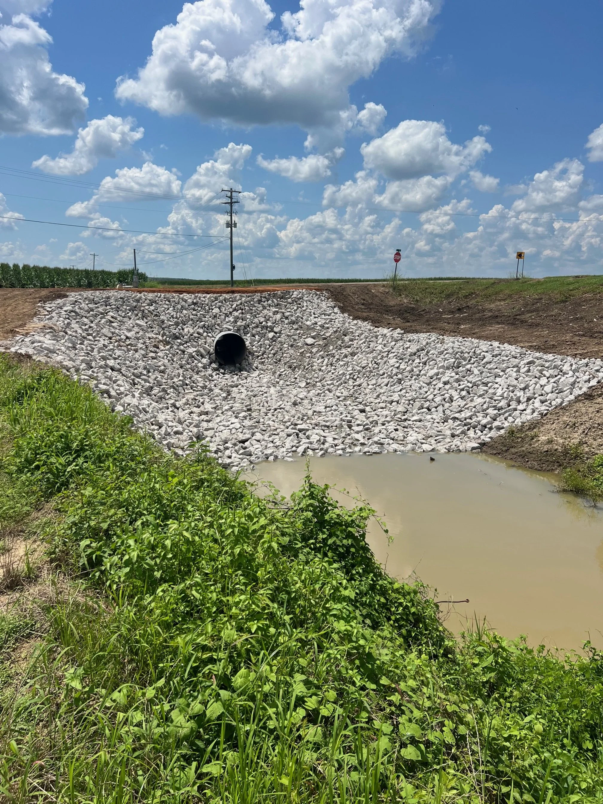 A culvert pipe installed in a rocky embankment near a waterway under a cloudy sky.