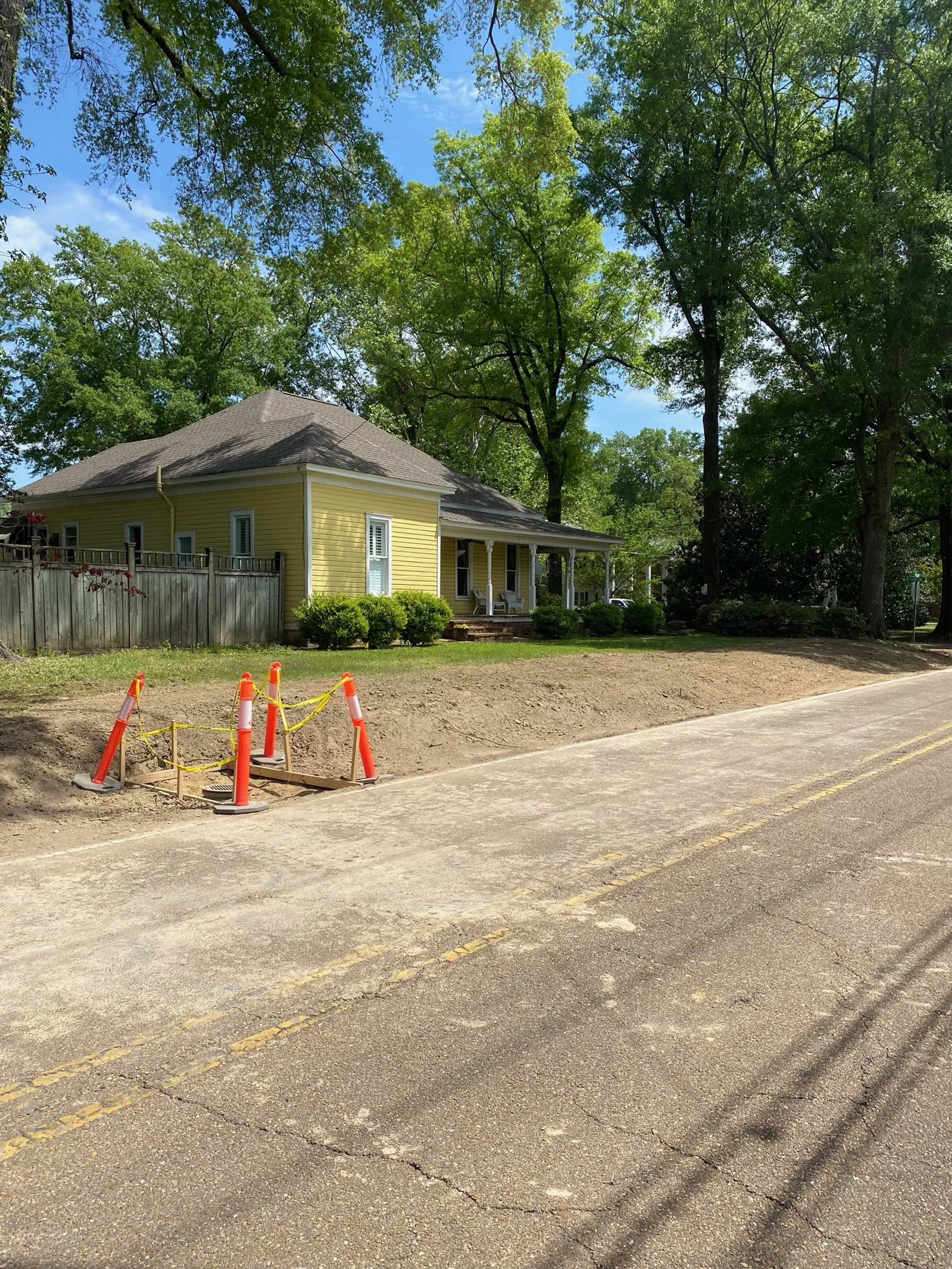 Street with construction cones and caution tape blocking a section of sidewalk, yellow house with front porch and trees in background.