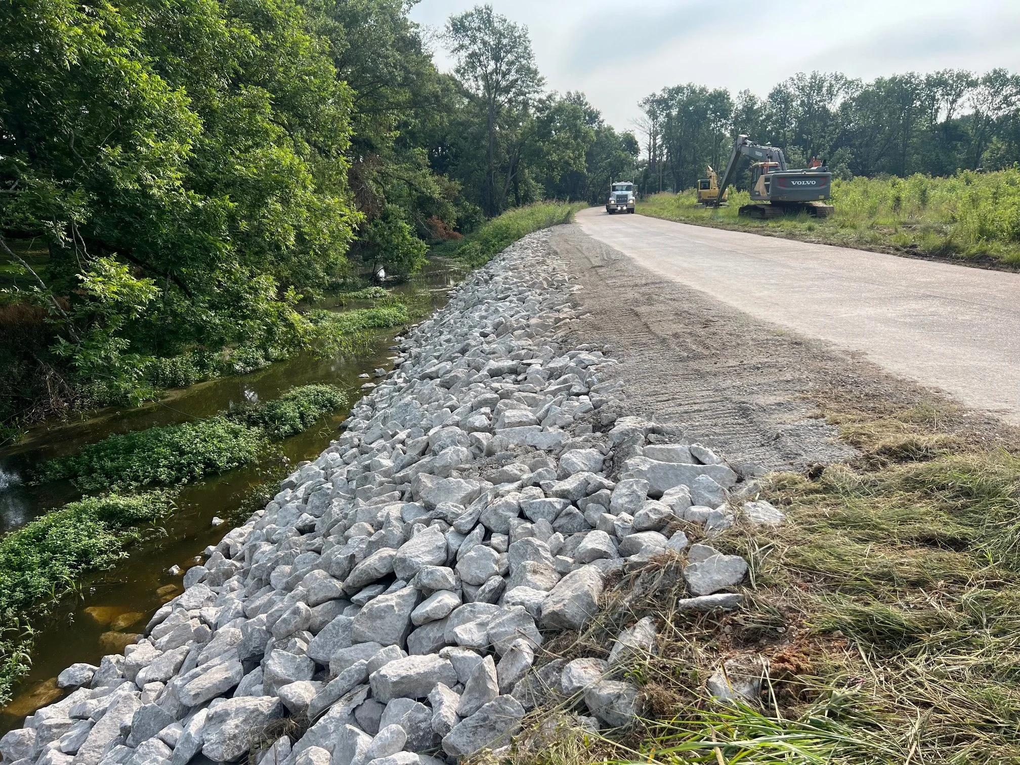 A construction site along a rural road with a gravel shoulder, heavy machinery, and a small creek bordered by green trees.
