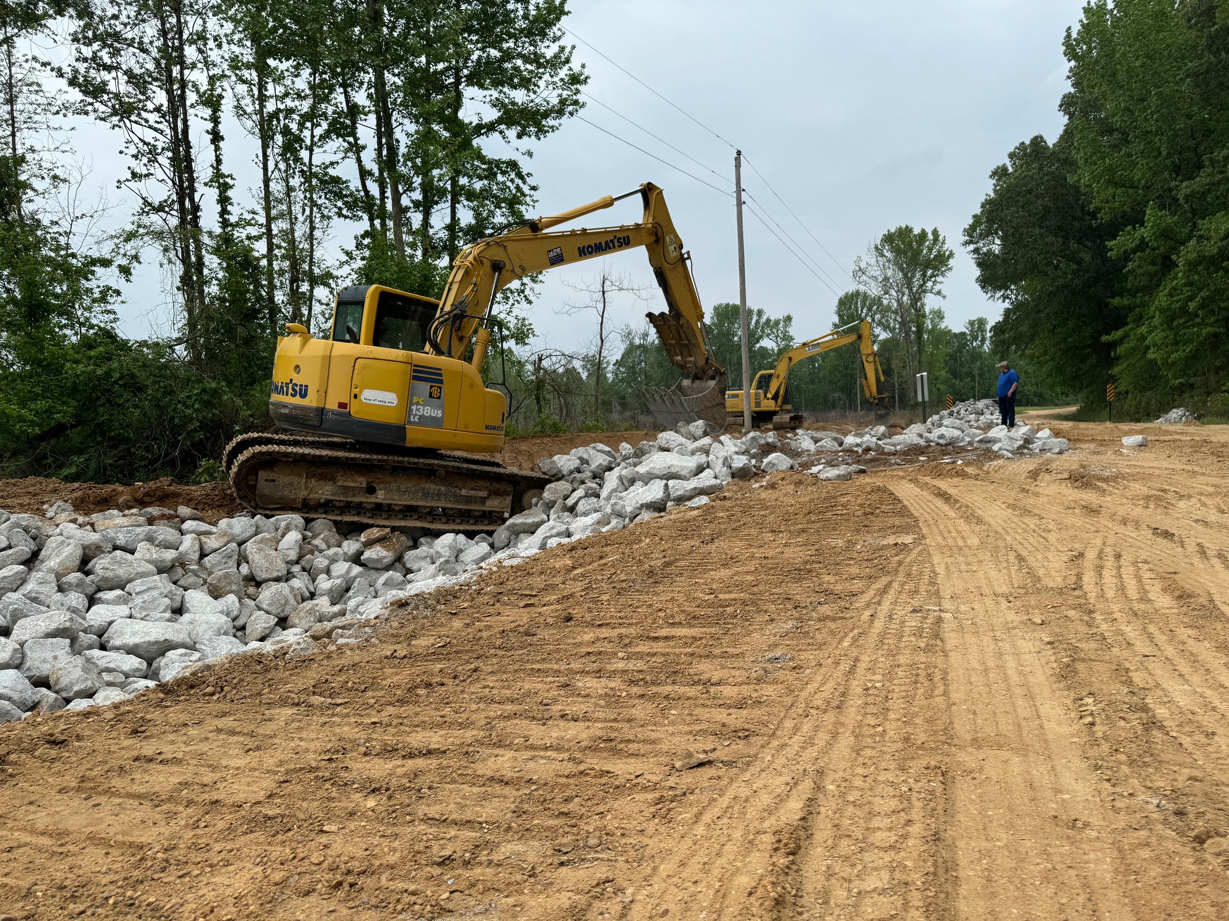 Two yellow excavators working on a dirt road, moving large rocks, with a person standing nearby, surrounded by green trees and power lines in an outdoor setting.