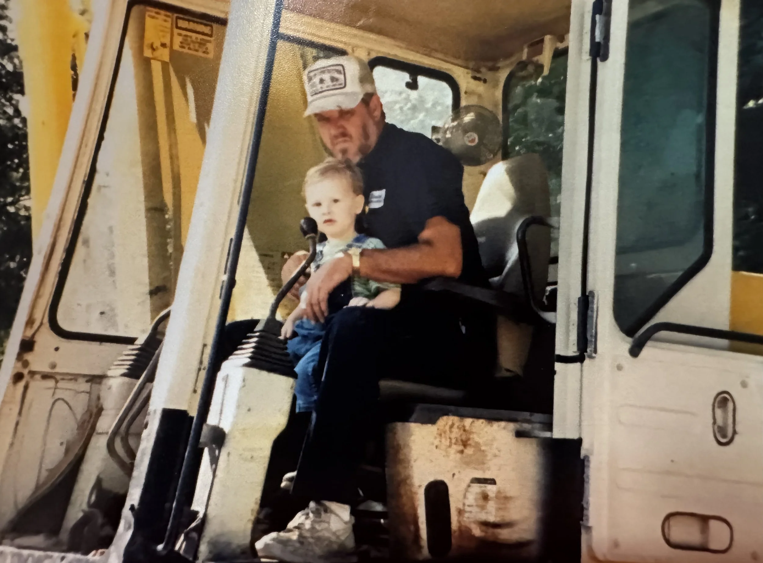 A man and a young boy sitting inside a construction vehicle, with the man holding the boy on his lap.
