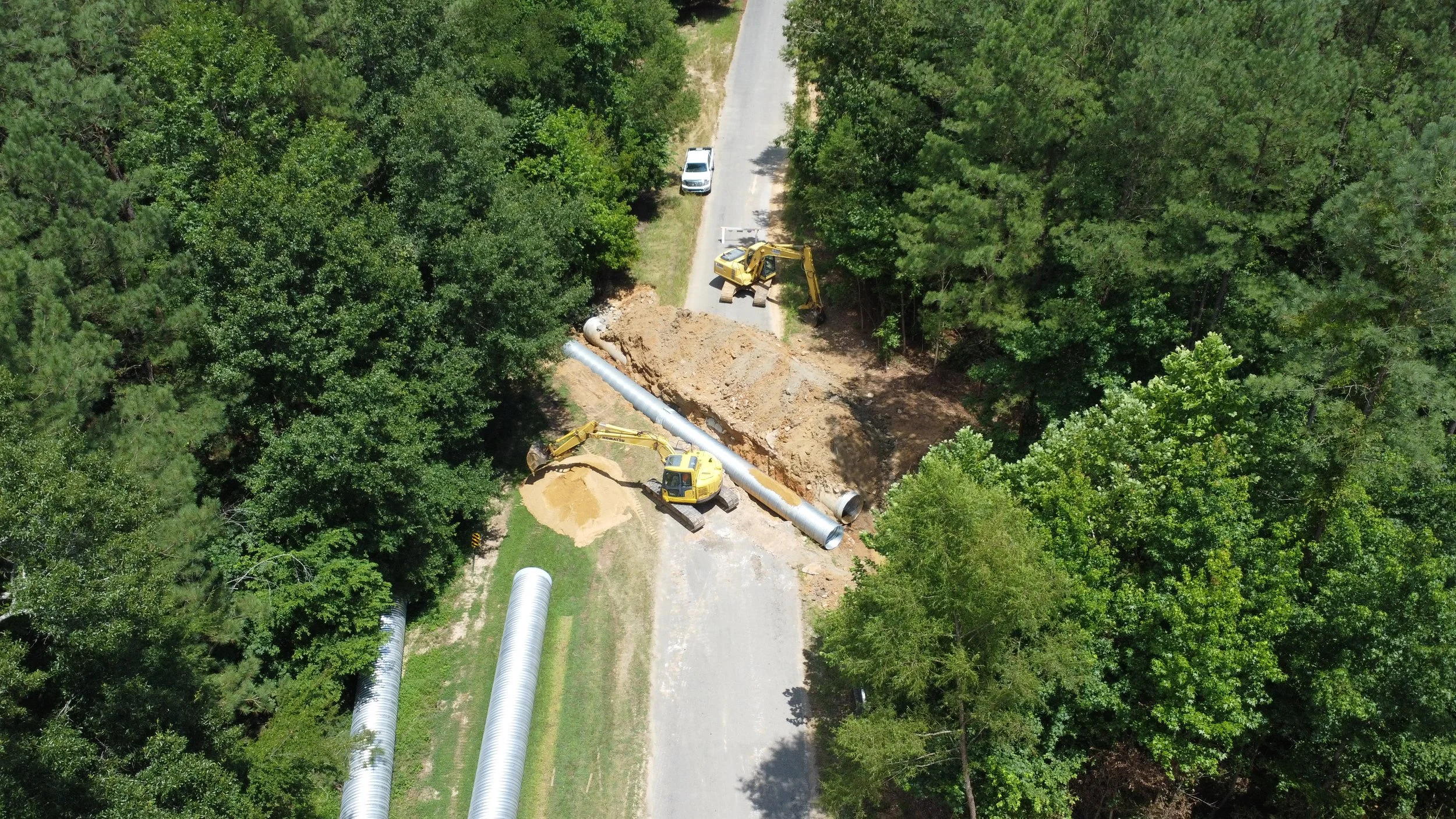 Aerial view of construction work where large pipes are being laid underground in a forested area, with excavators and a work truck present.