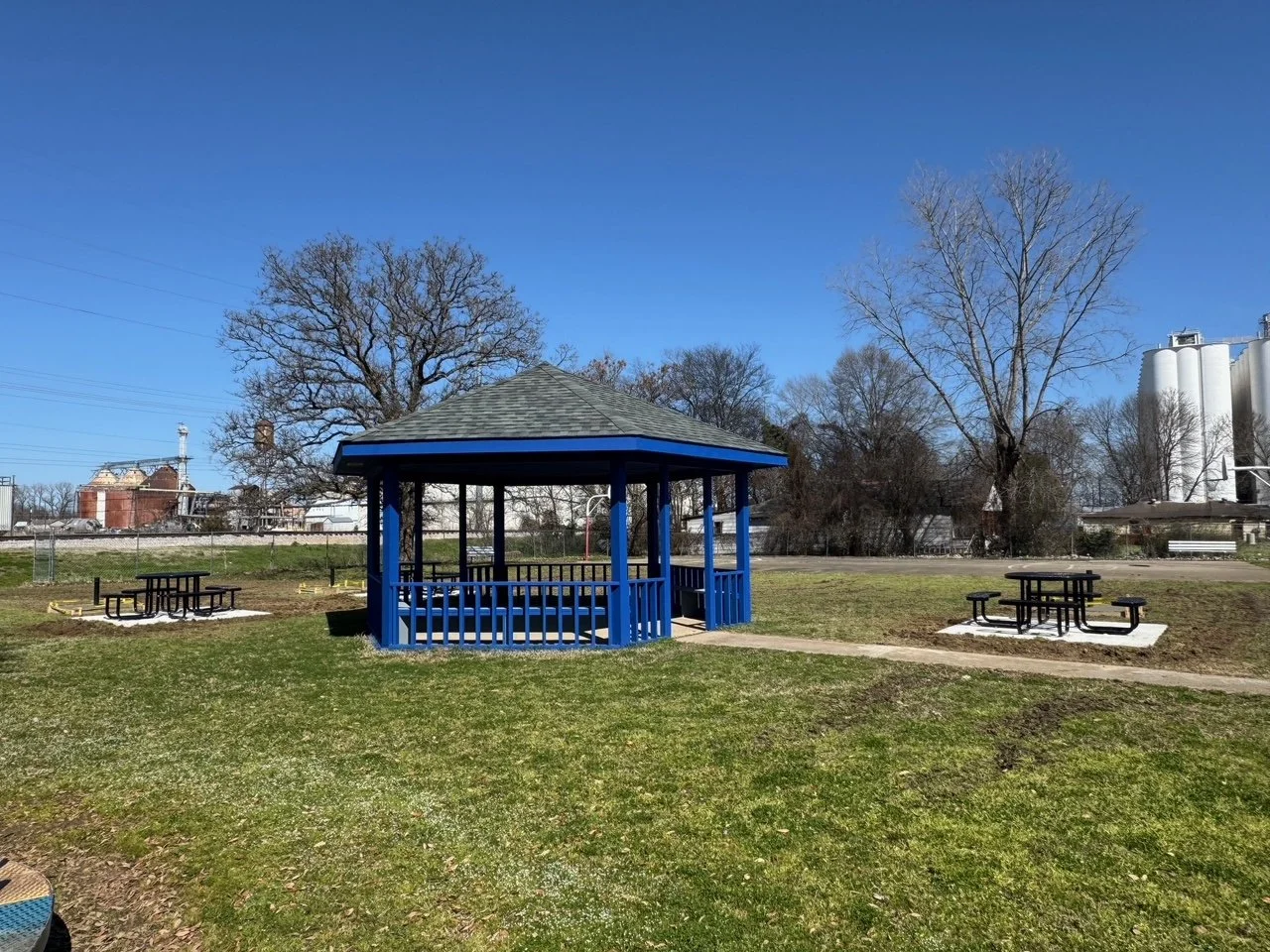 A park with a blue gazebo, two picnic tables, and leafless trees on a sunny day.