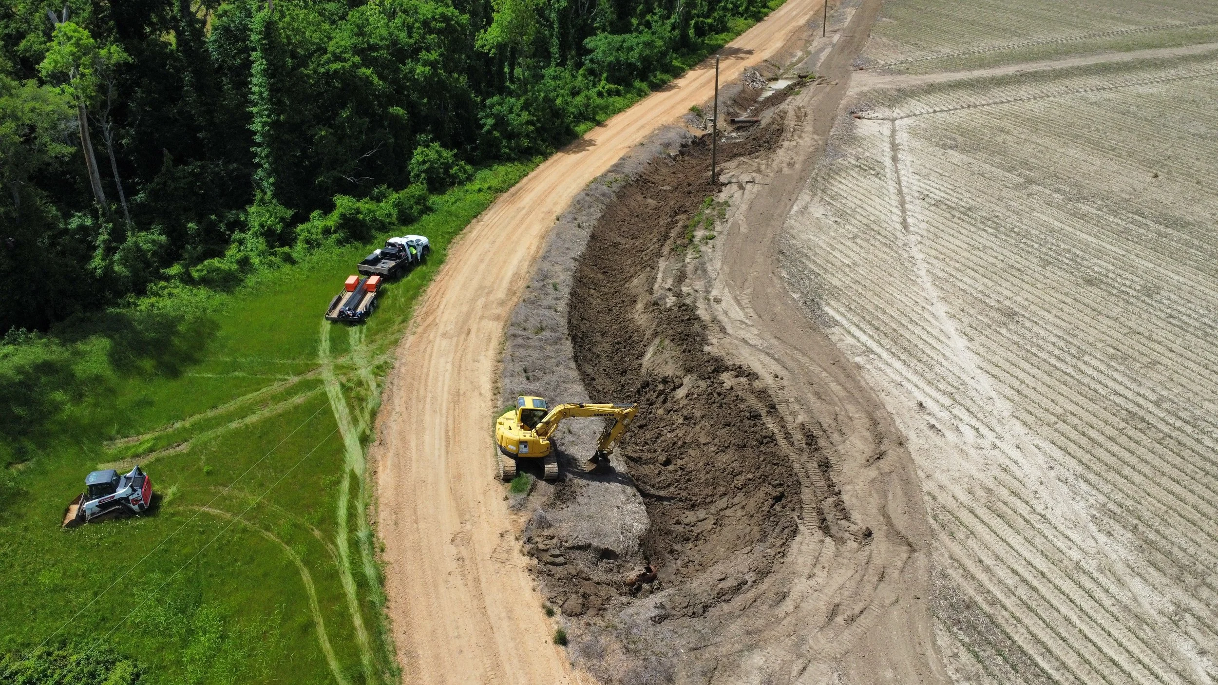 Construction site with excavator digging along a dirt road next to a field and a forest area.