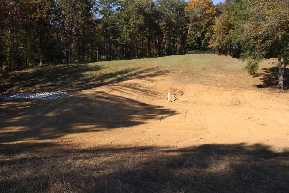 A cleared area of land with exposed dirt, surrounded by trees with fall foliage, and a small patch of snow on one side.