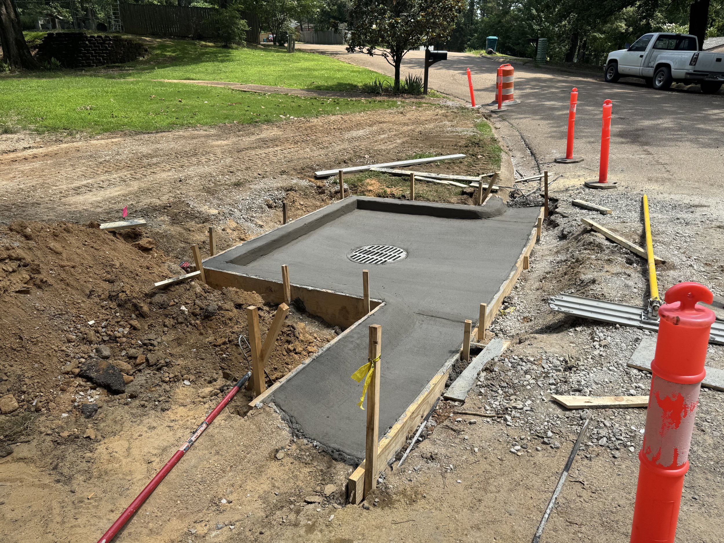 Construction site with freshly poured concrete sidewalk, orange and yellow safety cones, wooden forms, construction tools, and a storm drain in the center of the new sidewalk.