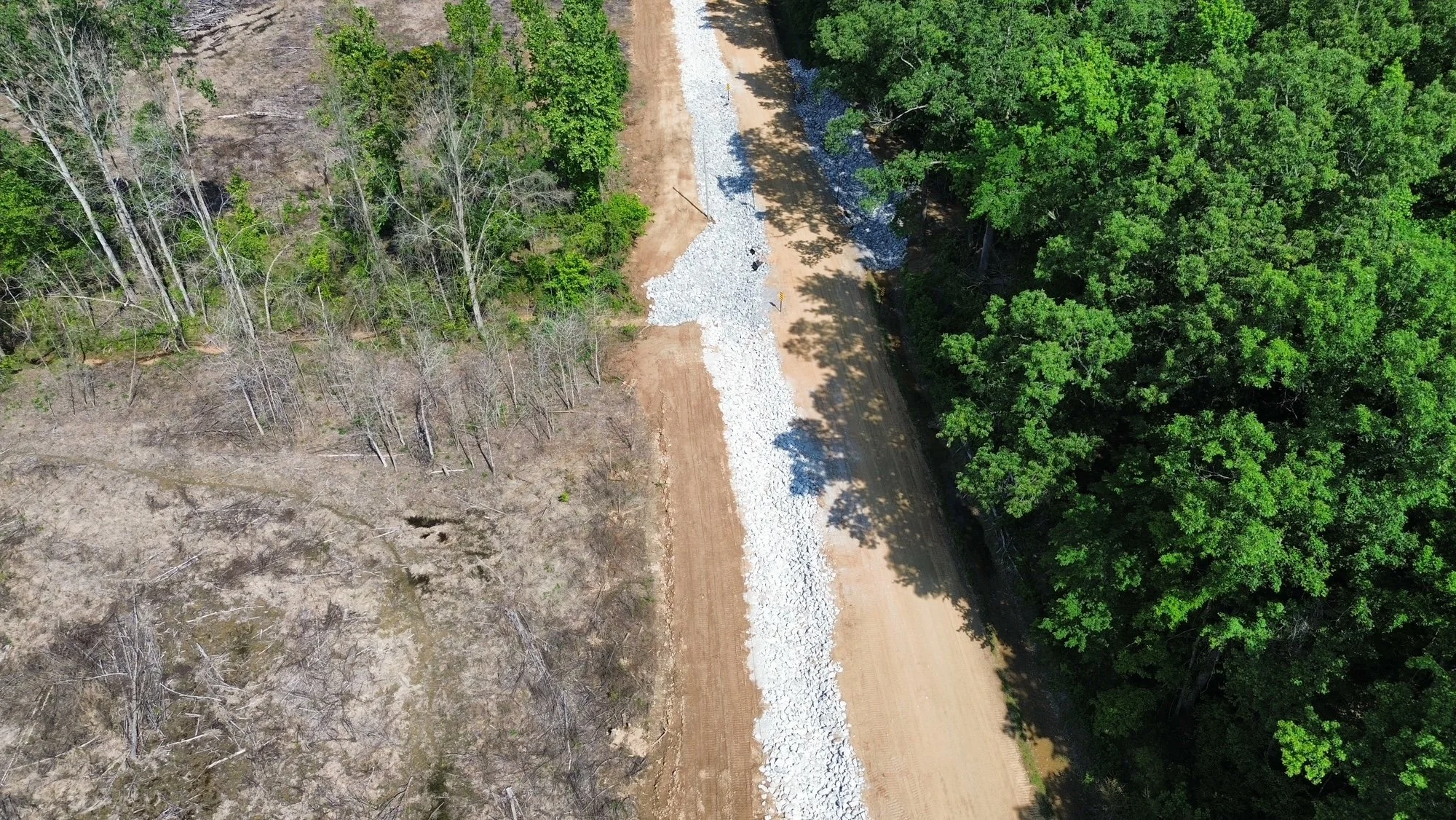 Aerial view of a dirt road under construction with gravel in the center, surrounded by deciduous trees on both sides, some with green leaves and others without leaves.