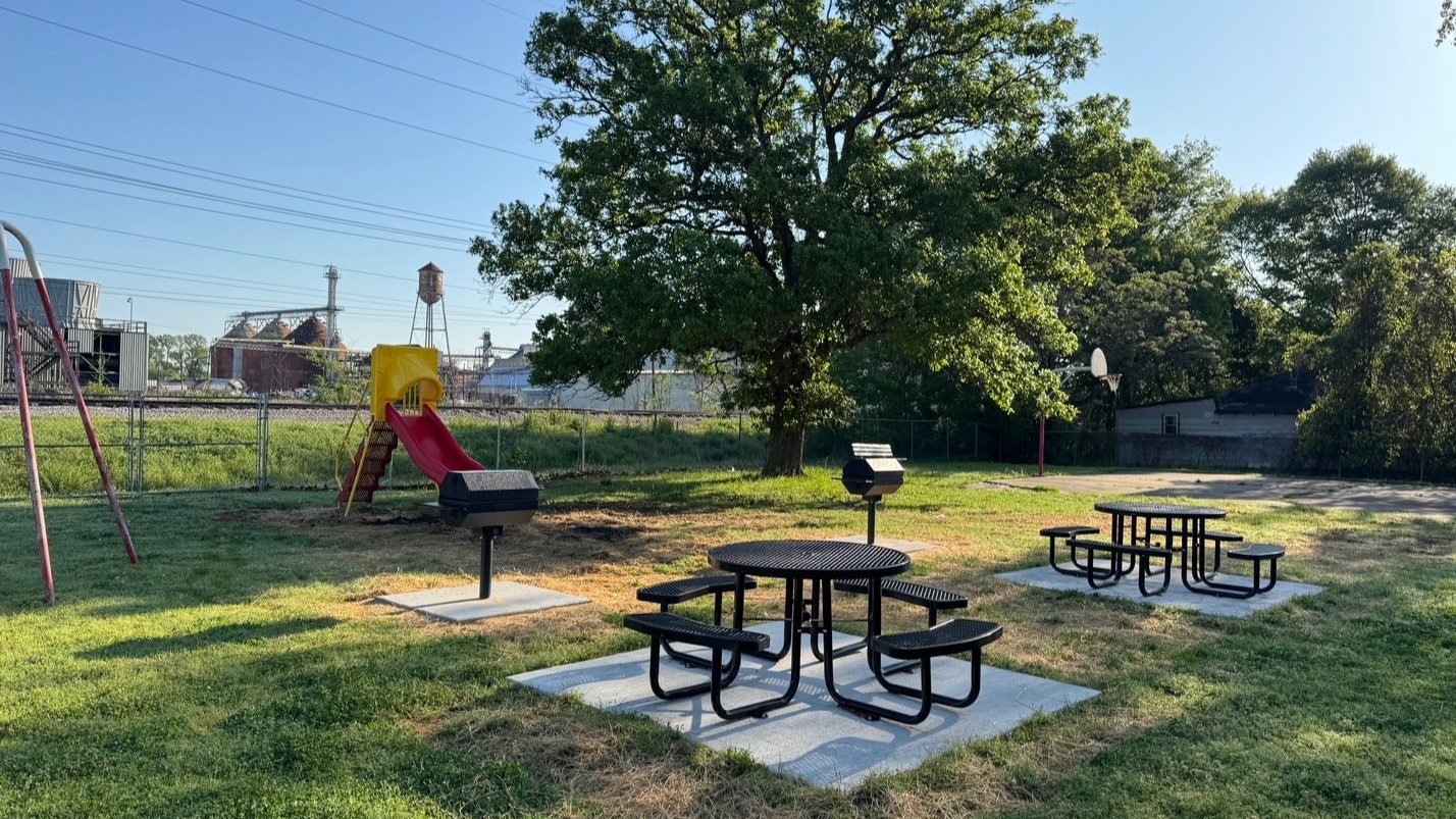Children's playground with slide, swings, picnic tables, and a basketball hoop, surrounded by grass, a large tree, and industrial structures in the background.