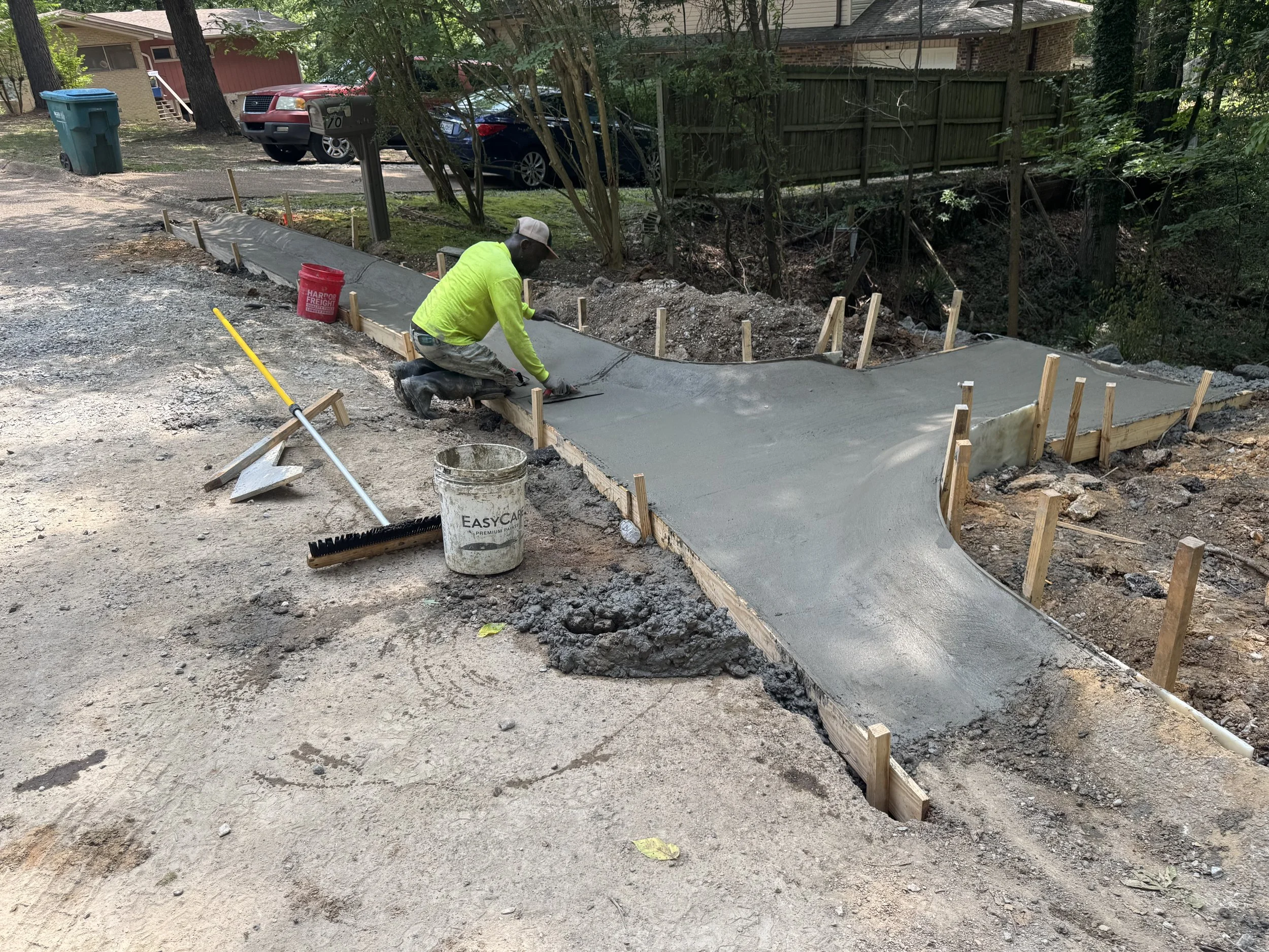 A construction worker in a neon green shirt and camouflage pants is kneeling on the ground, smoothing freshly poured concrete on a winding sidewalk path. Wooden forms outline the shape of the sidewalk, and tools, including a broom and a bucket, are nearby. In the background, there are parked cars, trees, a house, and a wooden fence.