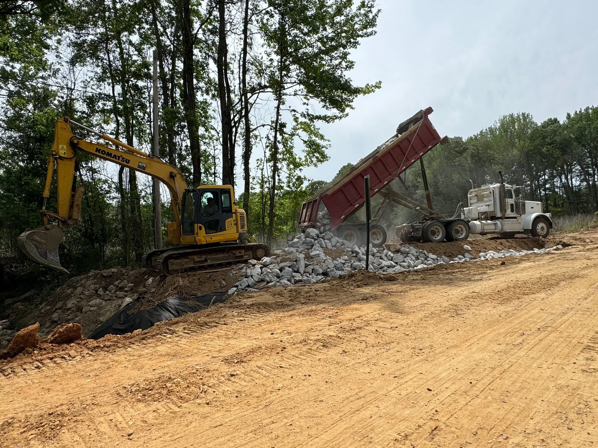 Construction site with a yellow Komatsu excavator and a white dump truck unloading rocks onto a dirt road surrounded by trees.