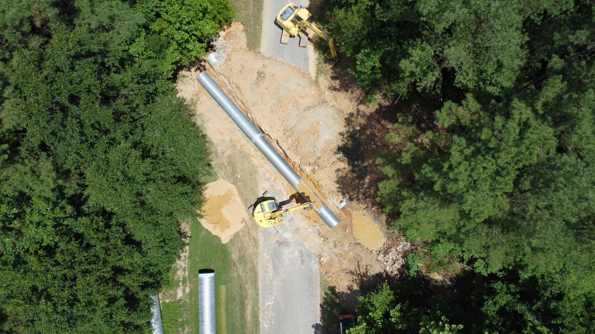 Aerial view of construction site with large metal pipe, excavators, and clearing area surrounded by dense trees.