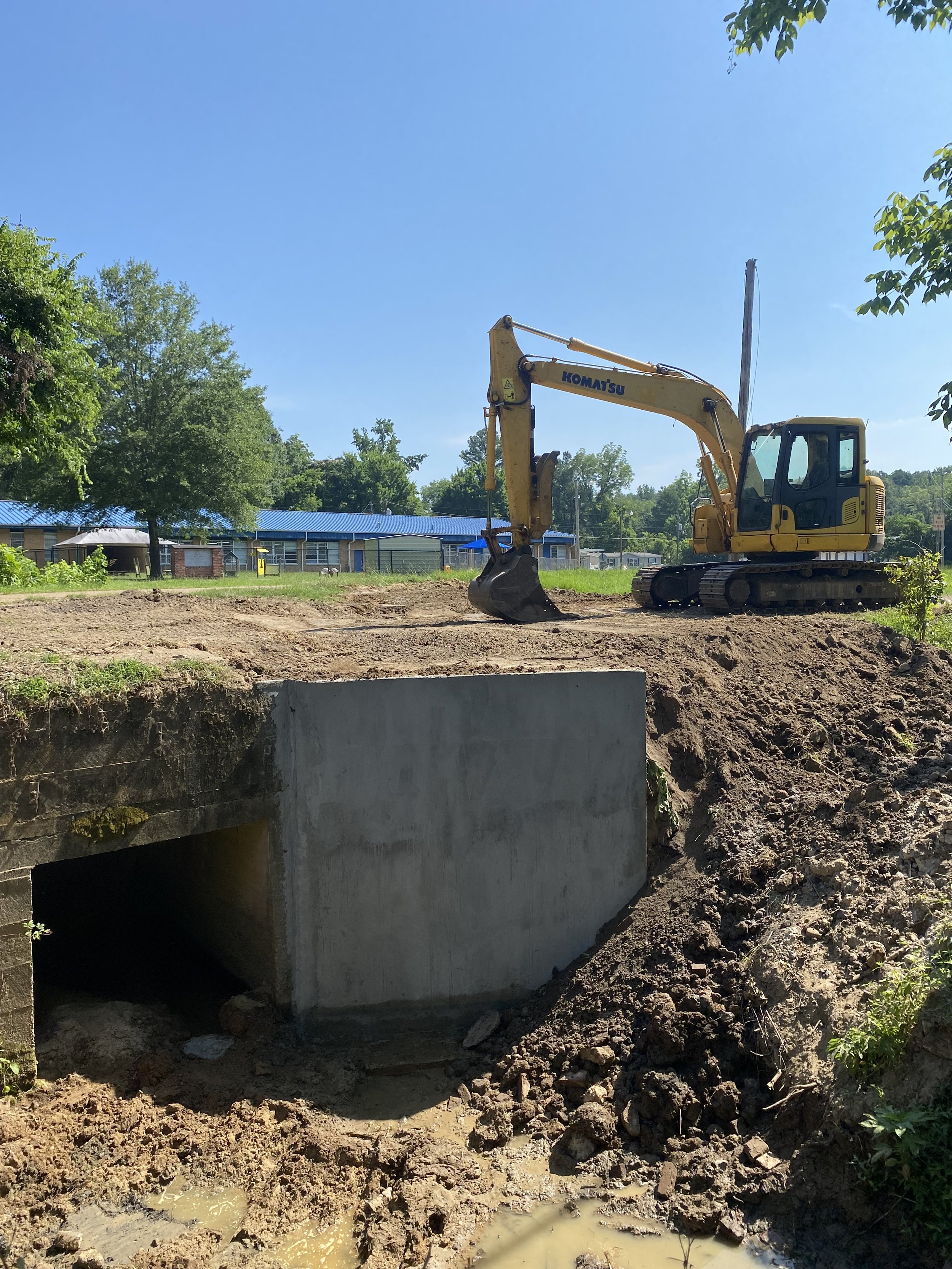 Construction site with a yellow Komatsu excavator positioned near a concrete culvert, dirt and mud in the foreground, and a blue sky with green trees and buildings in the background.