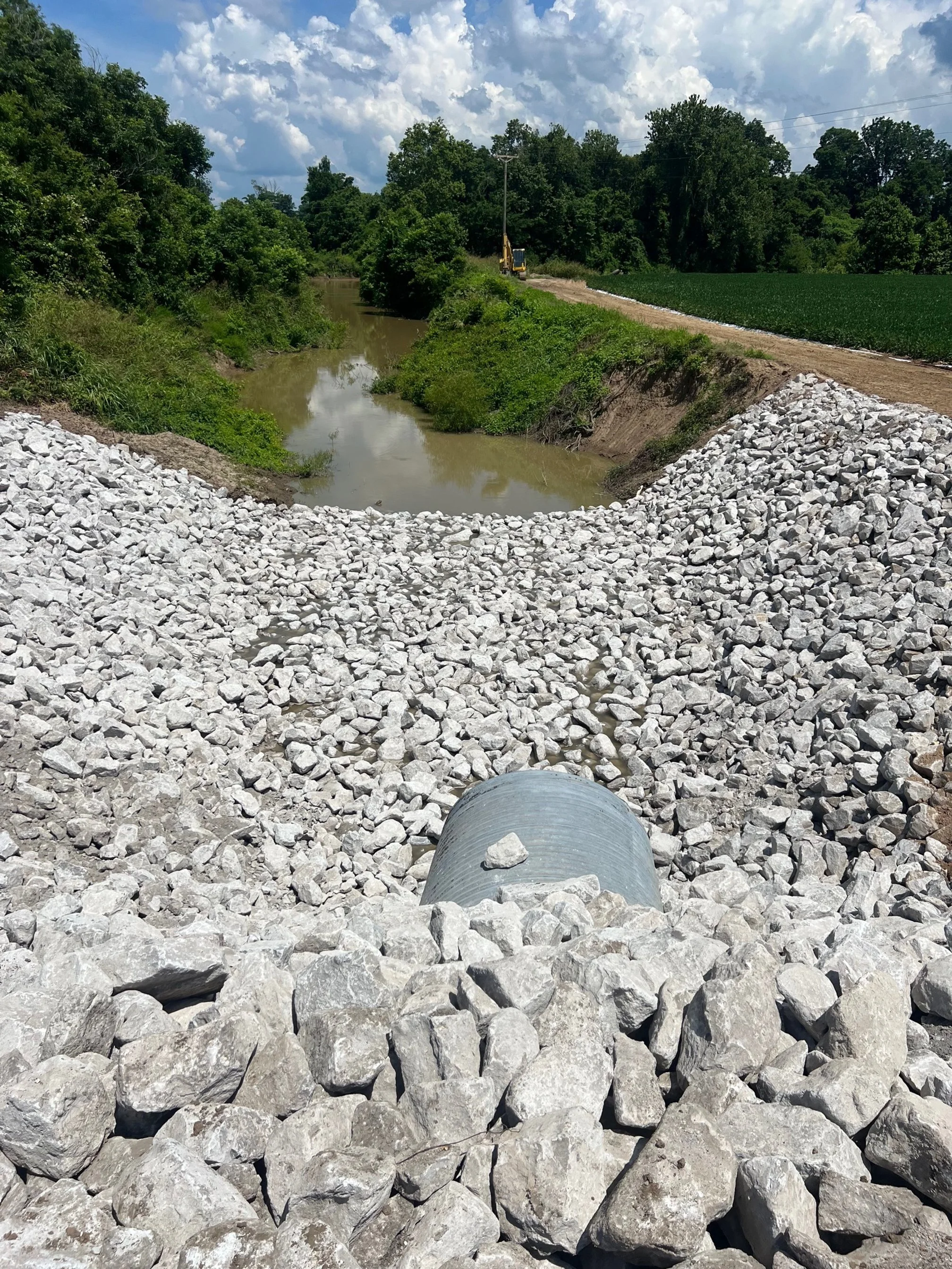 Construction site showing a pipe embedded in a bed of gravel, with a small river or pond in the background and trees lining the horizon on a partly cloudy day.
