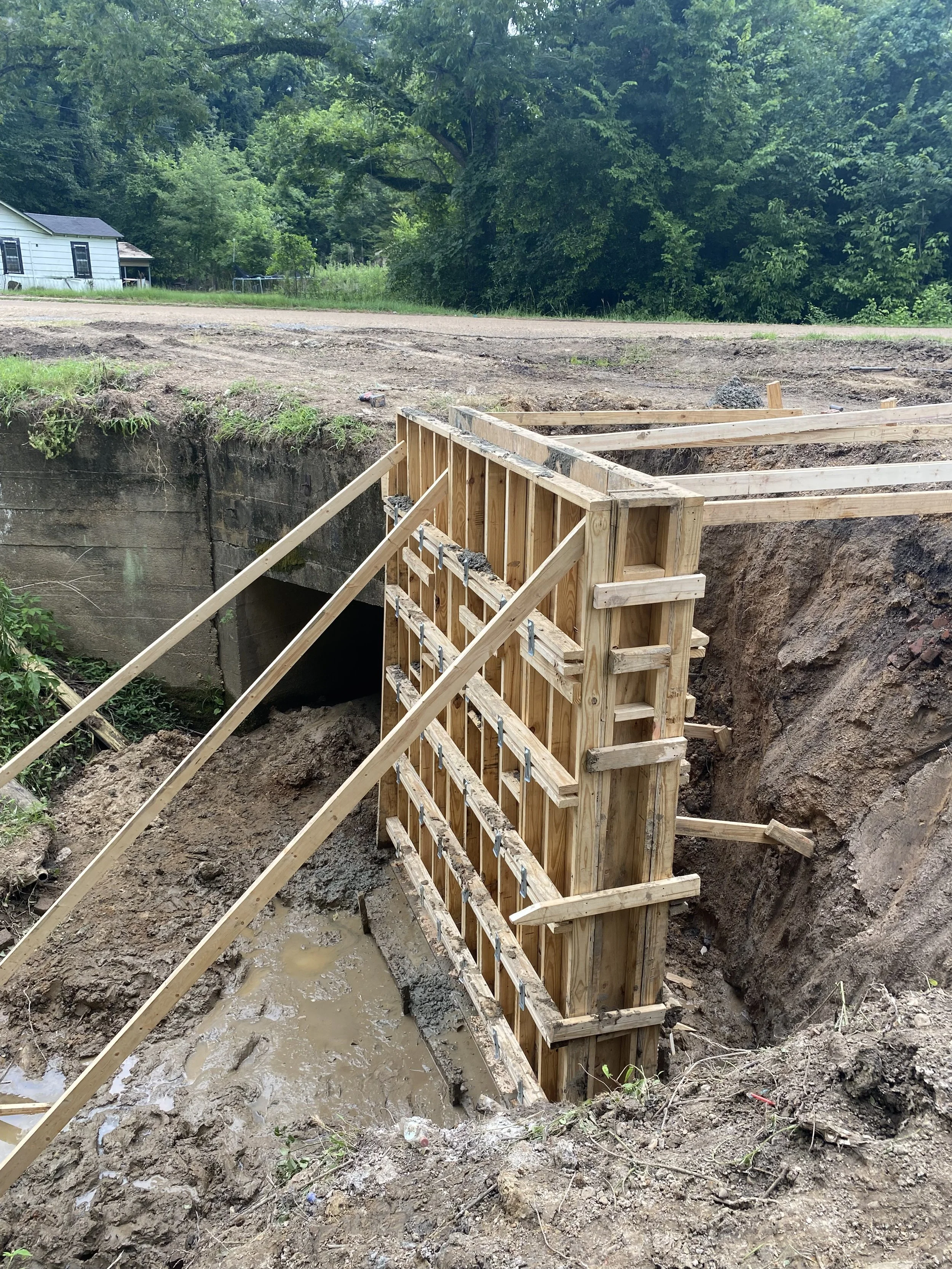 Wooden formwork for a construction project built on an excavated area near a small tunnel, with muddy ground and construction supports in a rural setting.