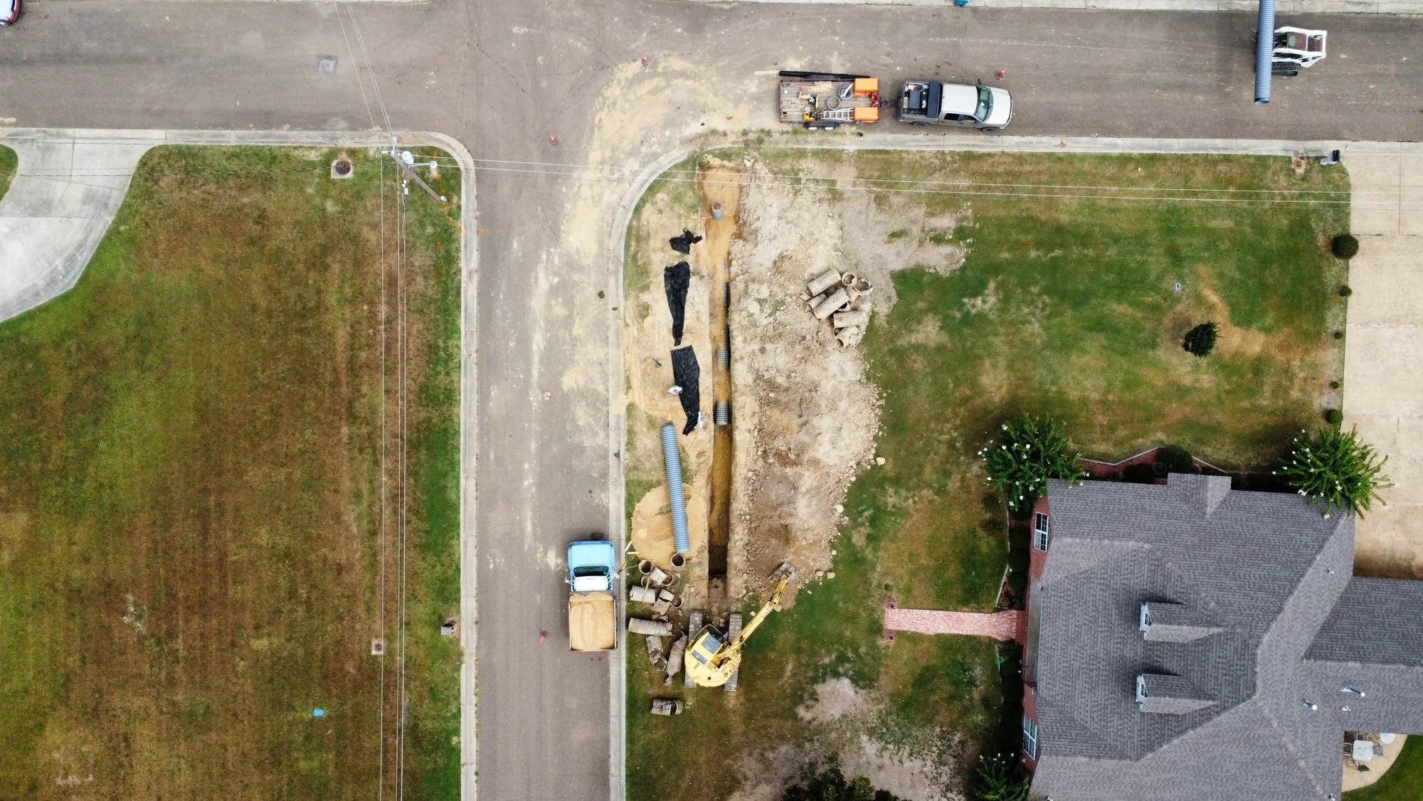 Aerial view of a backyard with ongoing construction work, including an excavator, construction materials, and a trench, adjacent to a house and a street with vehicles.