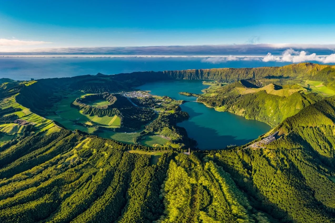Aerial view of a volcanic caldera filled with a lake, surrounded by lush green hills and patches of farmland, with a small village and forests, under a blue sky with some clouds.