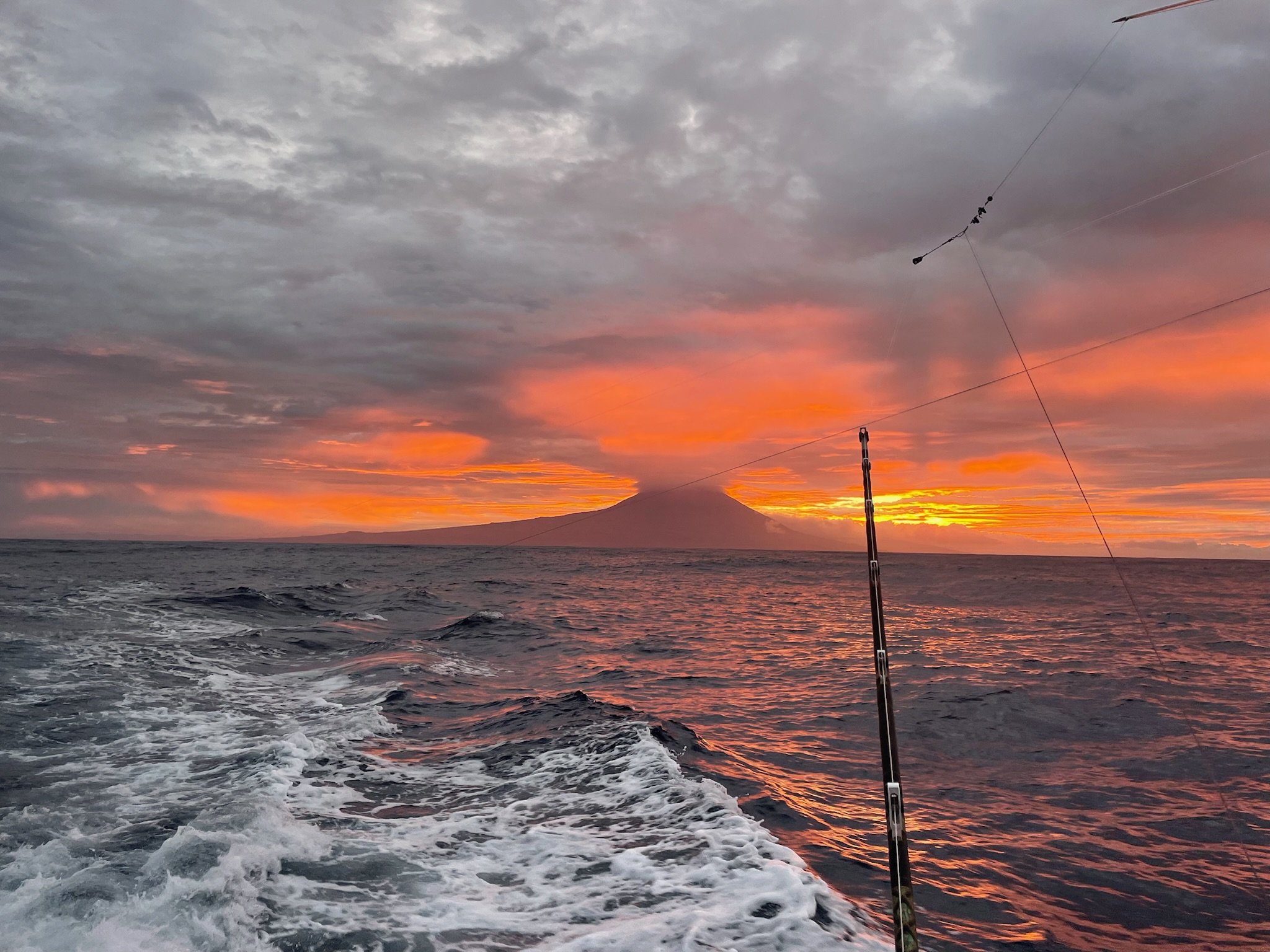View from a boat on the ocean during sunset with a fishing rod in the foreground and a volcanic mountain in the distance.