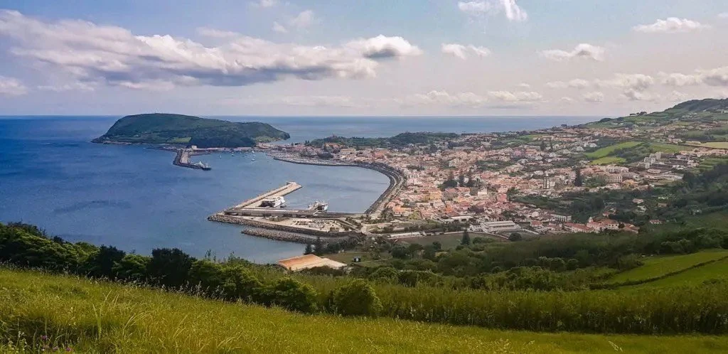 A panoramic view of a coastal city with a harbor, harbor structures, a bay, and surrounding green hills under a cloudy sky.